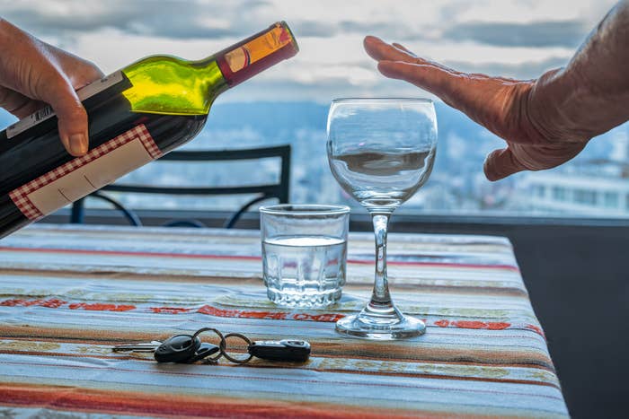 Person som nekter vin, gestikulerer «nei»; med hånden. Flaske og vinglass på bordet med bilnøkler og vannglass. Utendørs utsikt i bakgrunnen