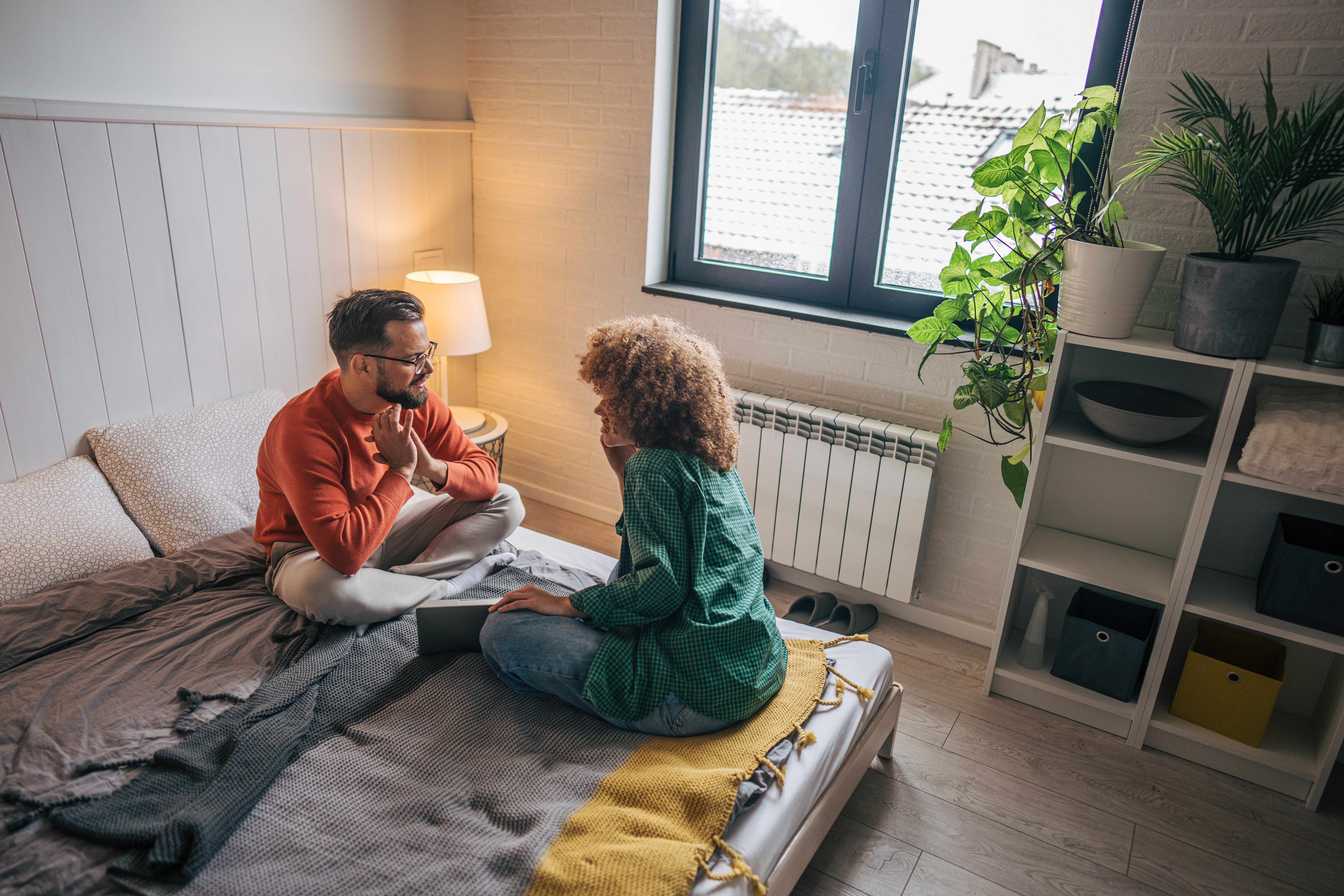 To personer sitter på en seng i et koselig rom, engasjert i en inderlig samtale, med myk belysning og skaper en intim atmosfære