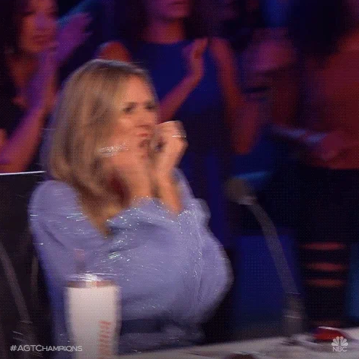 Person ved et talentshow -dommerbord, synlig begeistret, klappende hender og smilende, med et publikum som klapper i bakgrunnen