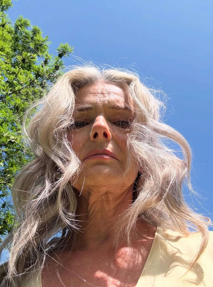 Person med bølget, grått hår ser ned på kameraet mot en klar himmel- og trebakgrunn, og formidler et kontemplativt humør