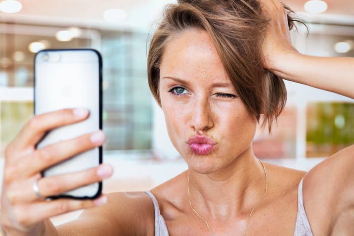 Person som tar en leken selfie, blinkende og puckerende lepper, med kort hår og uformell antrekk, i en lys setting