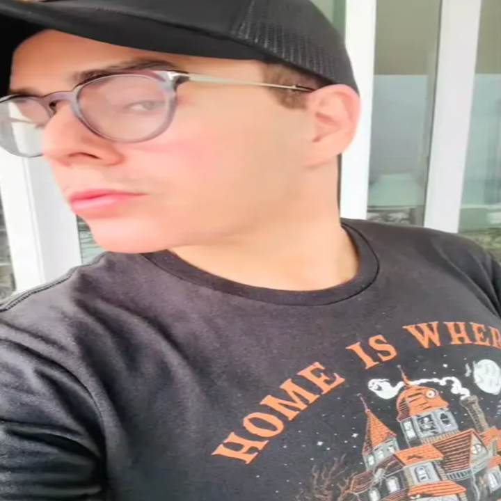 Person som har briller og en svart hette, ser på siden. De har en t-skjorte med en grafikk av et hus og tekst "Hjem er hvor"