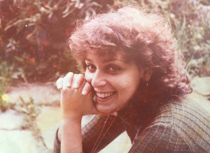 Person som smiler utendørs med krøllete hår, iført en strukturert, langermet topp, hvilende hender nær ansiktet