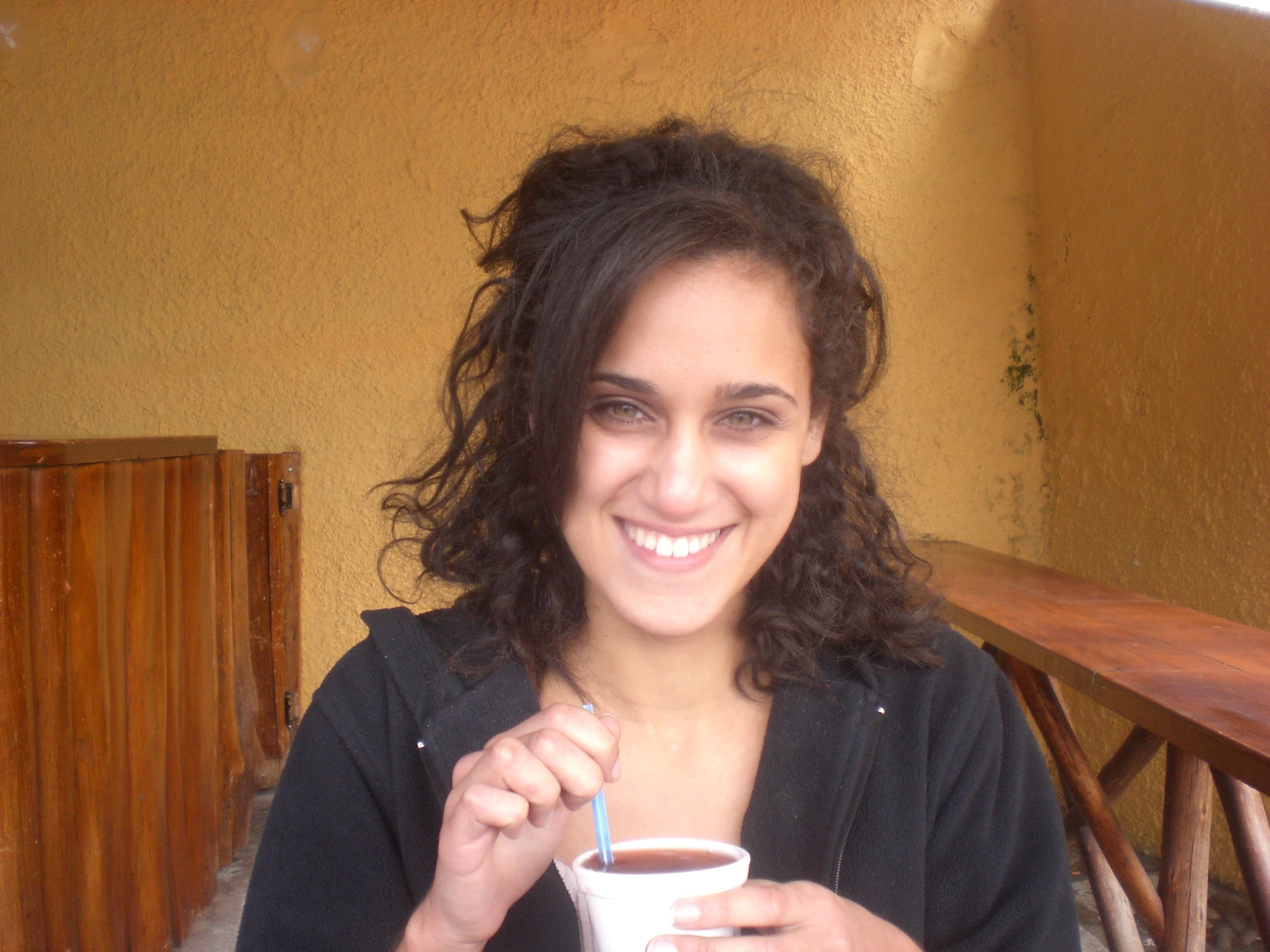 Personen smiler, holder en drink med et sugerør i tilfeldige omgivelser, iført en glidelås hettegenser