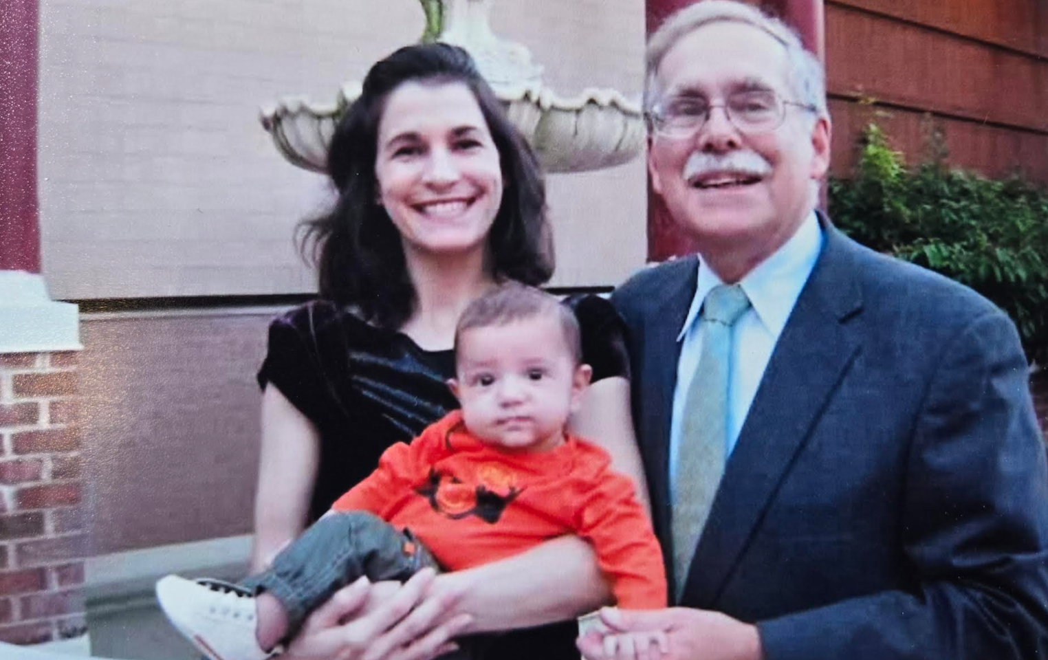 En kvinne og en eldre mann smiler mens han holder en baby foran en dekorativ fontene