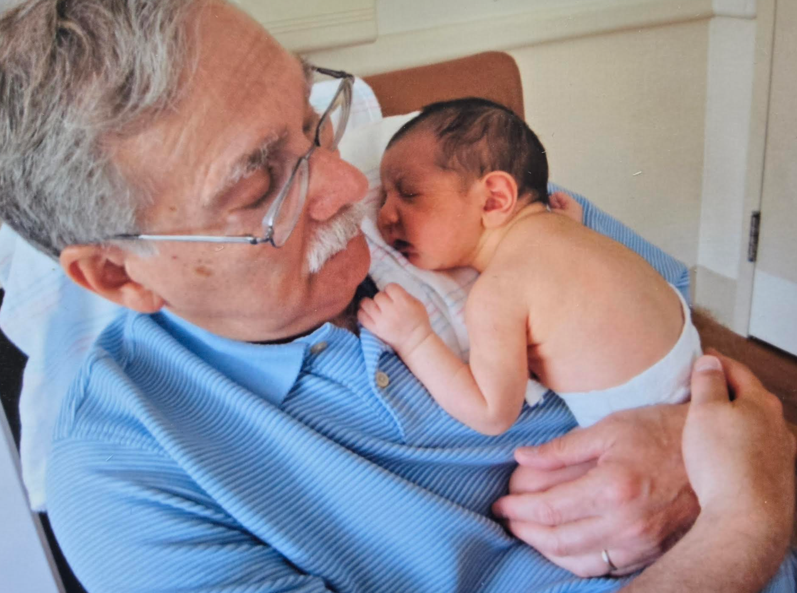 Eldre mann med briller som holder en sovende baby på brystet, begge virker fredelig og innhold