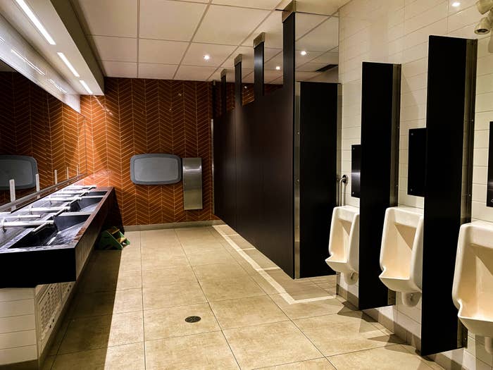 Moderne offentlig toalett med vasker, urinaler og avlukker synlige. Varm tre-lignende vegg aksenter gir stil til det rene, funksjonelle rommet