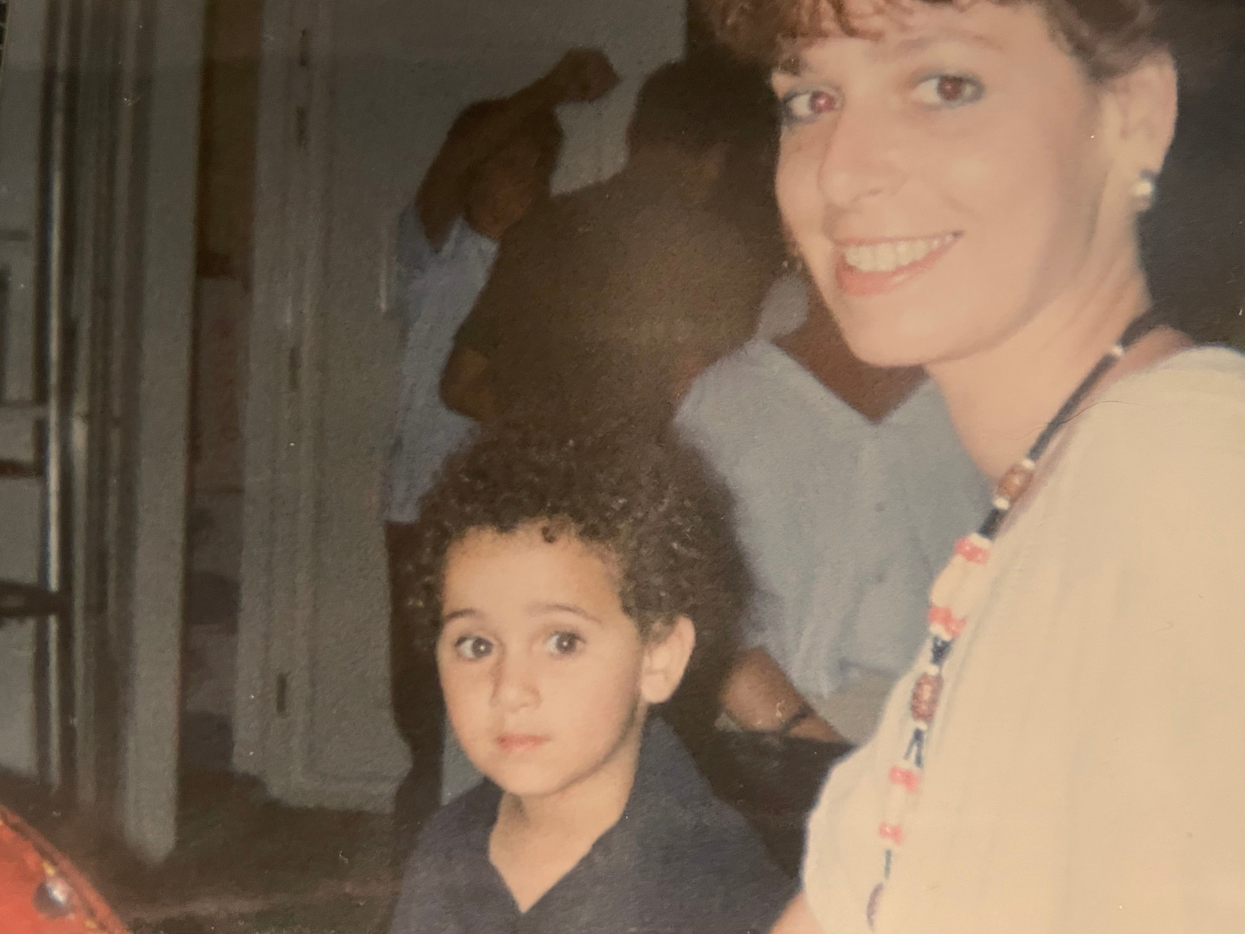 En kvinne smiler til kameraet mens et lite barn med krøllete hår ser litt på siden, sittende innendørs