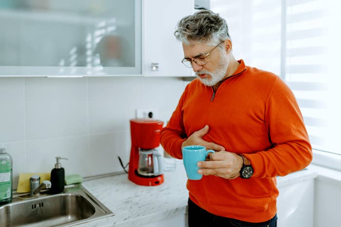 Mann på et kjøkken som holder et krus, virker gjennomtenkt, med en hånd på magen ved siden av en kaffetrakter og vask