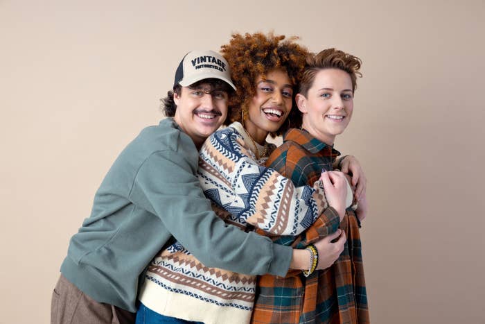 Tre venner smiler og poserer sammen. Den ene har en hette og genser, en annen har krøllete hår og en mønstret kofta, den tredje har en rutete skjorte