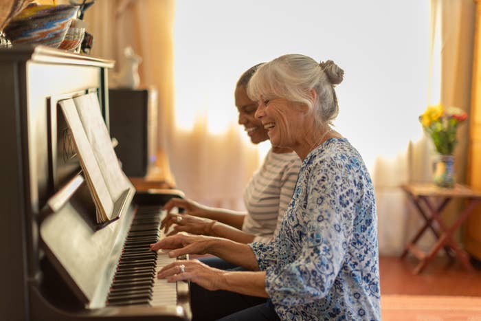 To kvinner, en eldre og en yngre, spiller gledelig piano sammen i et varmt opplyst rom, og deler et øyeblikk av musikk og latter