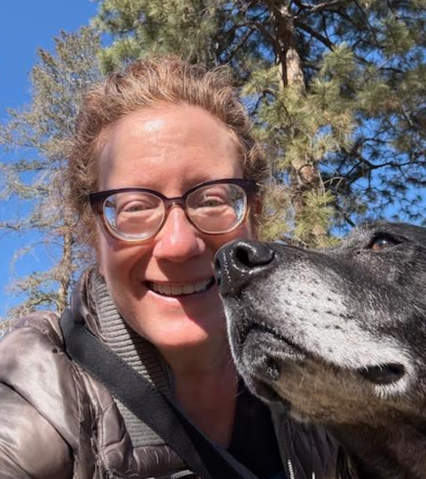 Personen smiler ved siden av en stor hund utendørs, med trær i bakgrunnen. Personen har briller og en puffy jakke