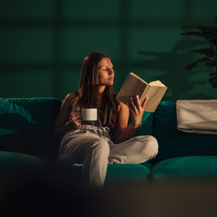 Person som slapper av på en sofa, holder et krus og leser en bok, med en plante i bakgrunnen