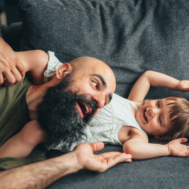 En skjeggete mann spiller gledelig med et smilende barn i en sofa, både ser avslappet og lykkelig