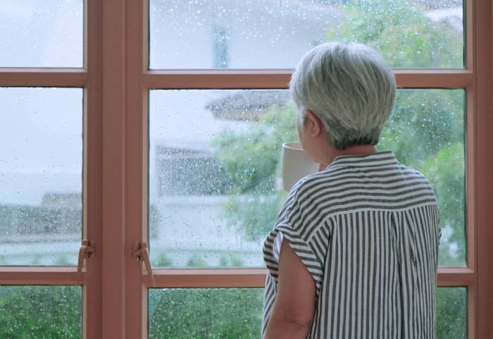 En person med kort, grått hår, iført en stripet skjorte, ser ut et regnprisede vindu og holder et krus