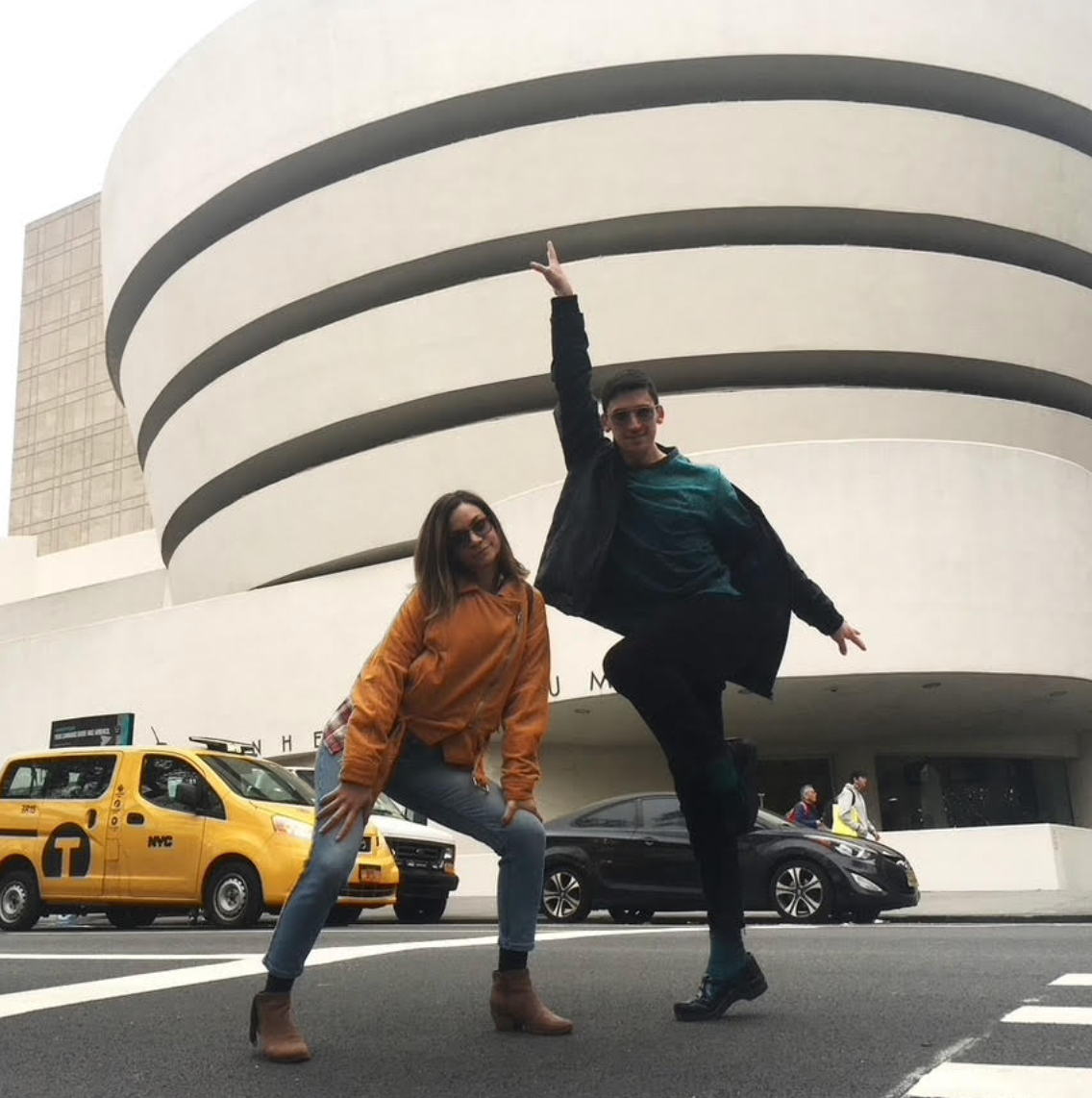 To personer poserer lekent på en bygate med Guggenheim -museet i bakgrunnen og drosjer som går forbi
