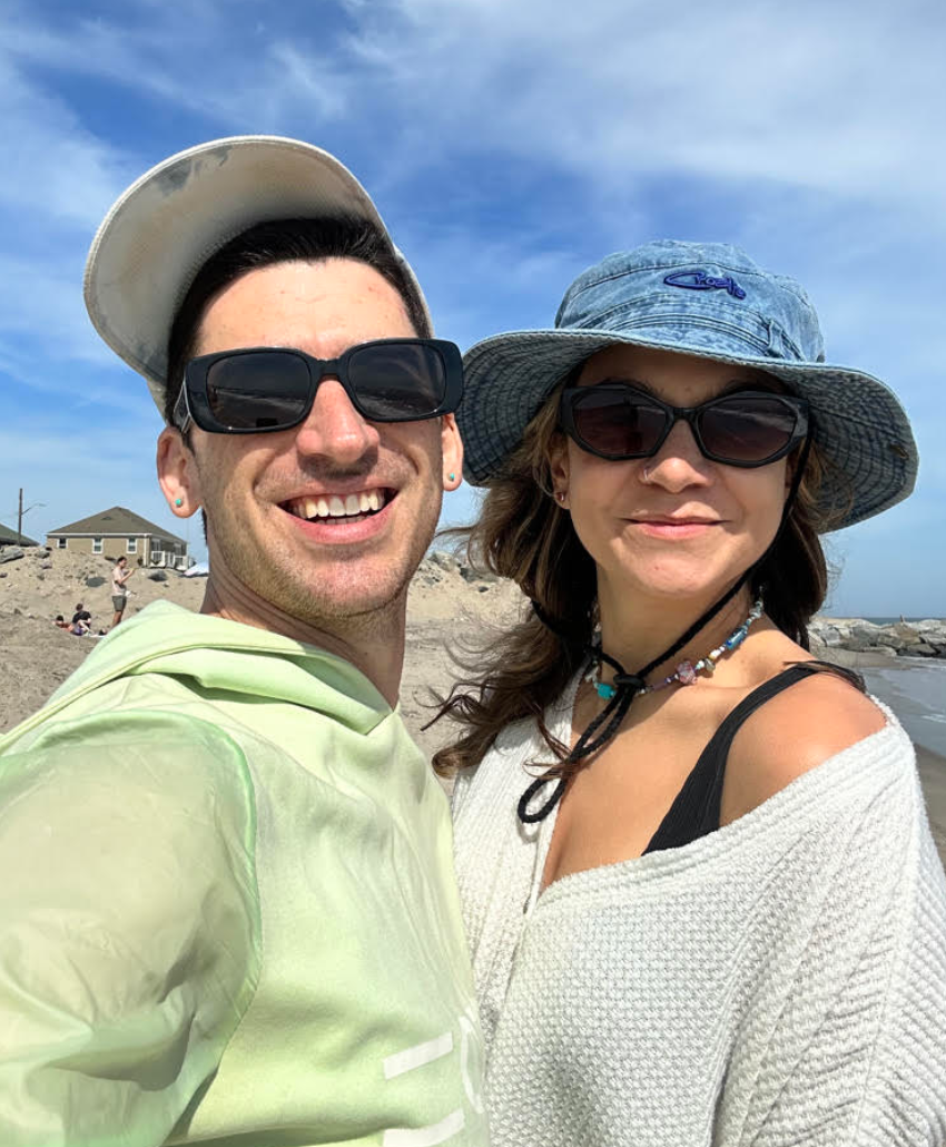 To personer som smiler på en strand. Begge har på seg solhatter og solbriller, og nyter en solskinnsdag