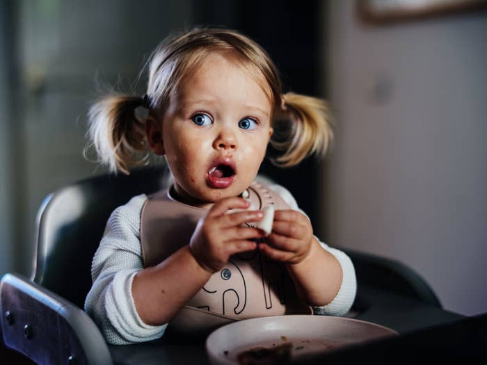 En pjokk med pigtails sitter i en høy stol og ser overrasket ut mens han holder mat