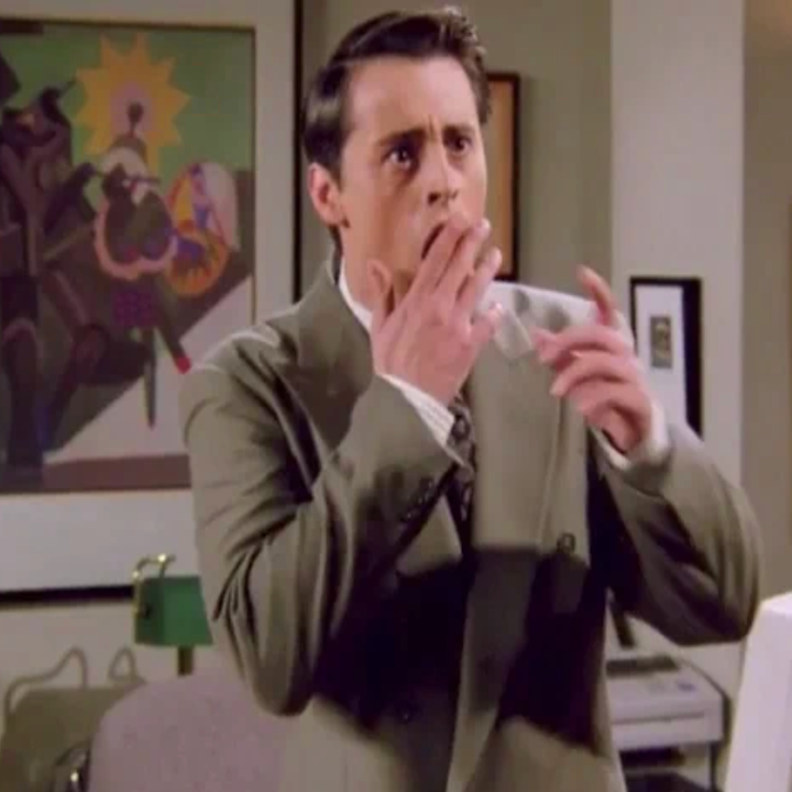 Person i en drakt som ser overrasket ut, hånddekker munnen, står i et rom med kunst på veggen