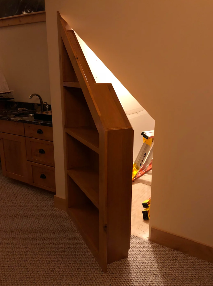 Skjult dør designet som en bokhylle, noe som fører til et lagringsområde under trapper. Rommet har en kjøkkenkrok med vask og skap