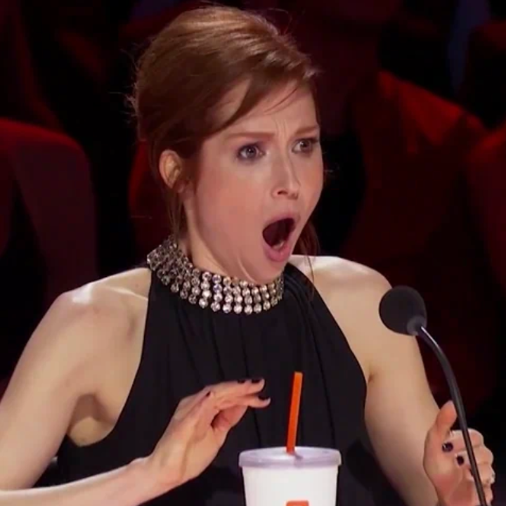 En sjokkert kvinne i en dommerpanel, iført en ermeløs topp med en perlet utringning, reagerer med brede øyne og en åpen munn