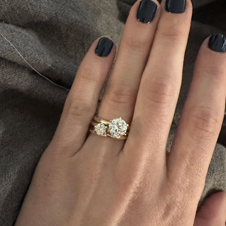 En hånd med mørk neglelakk viser en gullring med en stor rundkuttet diamant på en strukturert stoffbakgrunn