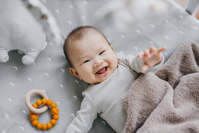En smilende baby som ligger på et teppe med et mykt leketøy og tenner ring i nærheten