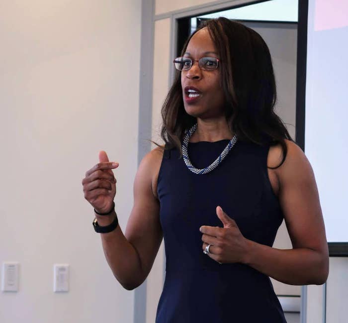 Person i en ermeløs kjole, snakker lidenskapelig, gestikulerer med den ene hånden, står i et rom med en skjerm i bakgrunnen