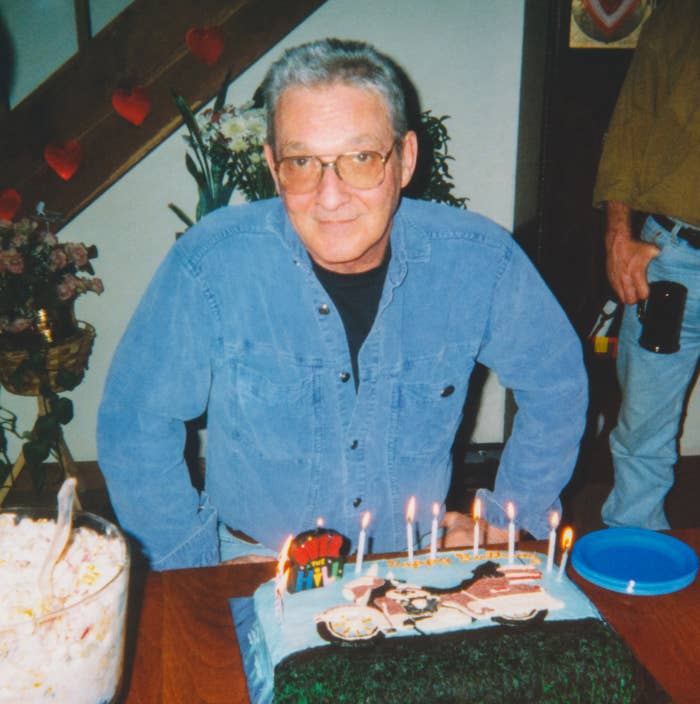 Mann i denimskjorta står ved en dekorert bursdagskake med tente lys innendørs