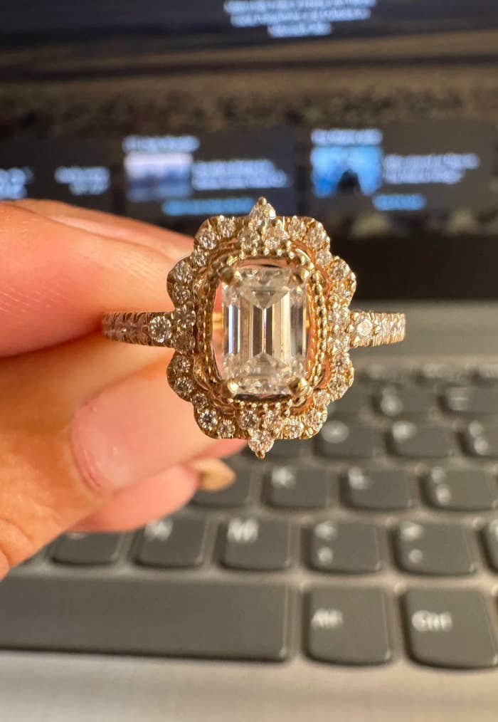 Et nærbilde av en hånd som holder en utsmykket diamantring med en rektangulær midtstein, satt mot en uskarp laptop tastaturbakgrunn