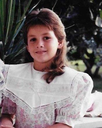 Barn som har på seg en blomstermønstret kjole med en stor hvit blonderkrage, smilende mykt, omgitt av grøntområder