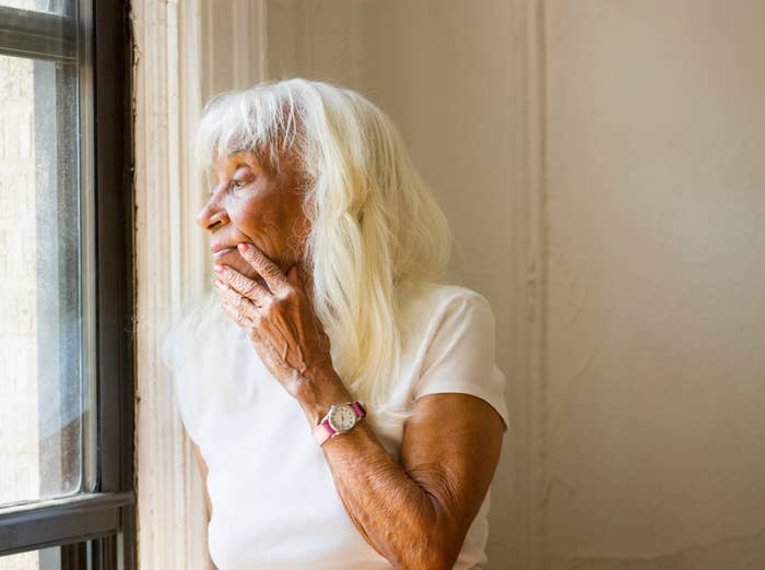 Eldre person med langt hvitt hår ser gjennomtenkt ut et vindu, hviler en hånd i ansiktet, iført en hvit topp og et armbåndsur