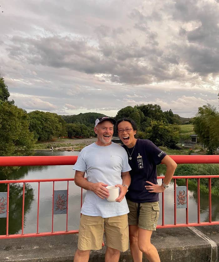 To personer som smiler på en bro ved en elv, en som holder en kaffekopp. Naturskjønn bakteppe med trær og en overskyet himmel