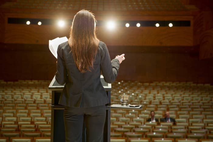 Person i forretningsantrekk snakker på et podium i et tomt auditorium med to personer som sitter i publikum