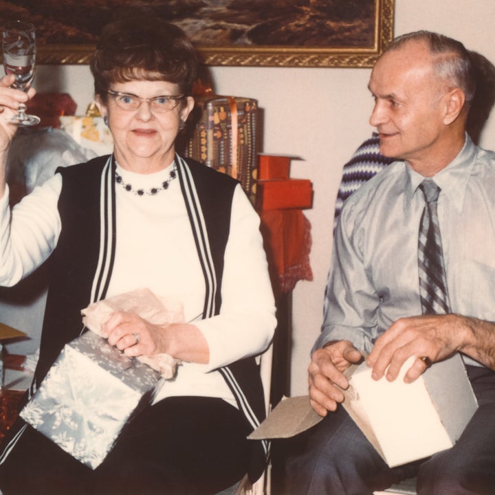 En eldre kvinne holder opp et glass, sitter ved siden av en eldre mann på en samling med gaver på bordet bak seg