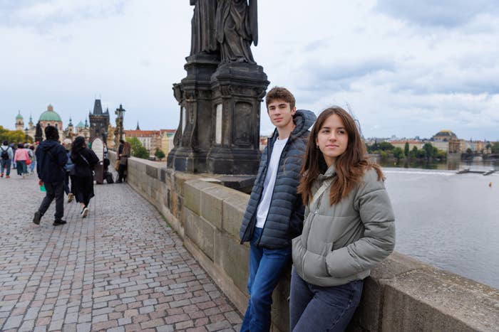 To personer i vinterjakker står på en brosteinsbro med statuer, med utsikt over en elv og bybildet i bakgrunnen