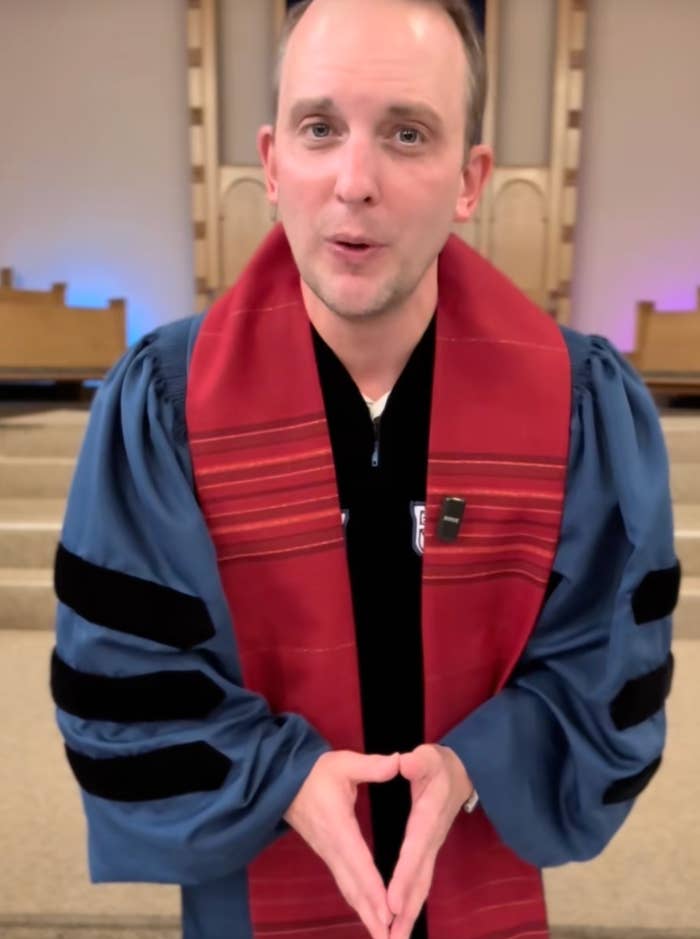 Person som har på seg en blå akademisk kjole med svarte fløyelsstriper og en rød stole, står innendørs og gestikulerer med hendene
