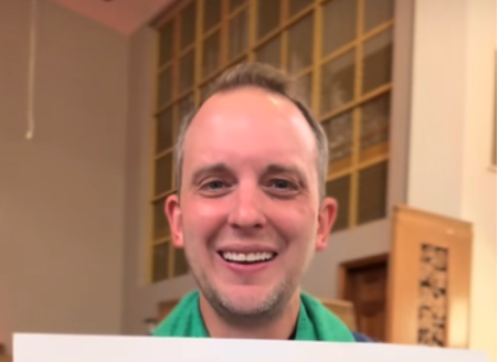 Person som smiler innendørs, holder en hvit rektangulær gjenstand under rammen. De har kort hår og er uformelt kledd