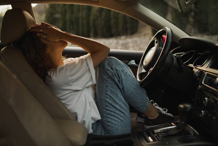 Person i uformelle jeans og t-skjorte som sitter i en bil, dekker ansiktet med hender, virker bekymret eller overveldet