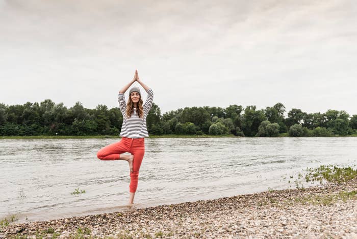 Person som praktiserer yoga i trepositur ved en elveside, stående på en rullesteinstrand med trær i bakgrunnen