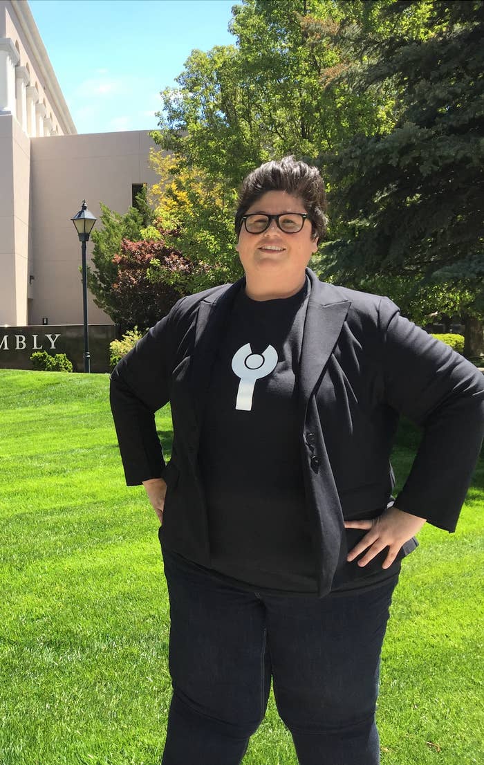 Person utendørs iført en tilfeldig svart blazer og T-skjorte med et sirkulært symbol, står selvsikkert på en solrik dag med trær og plen bak