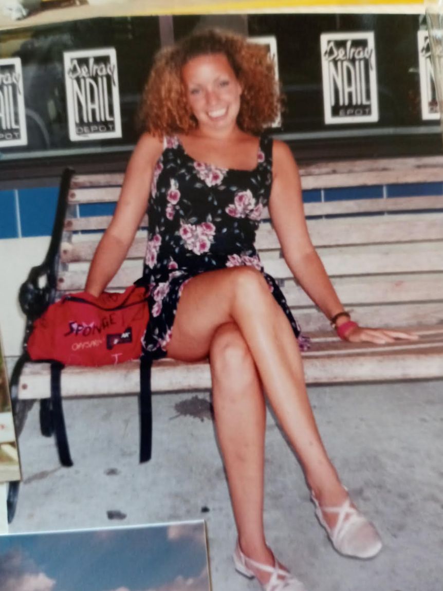 Kvinne sitter på en benk i en blomsterkjole og smiler. Bena er krysset, og hun har krøllete hår. En rød bag ligger på benken ved siden av henne