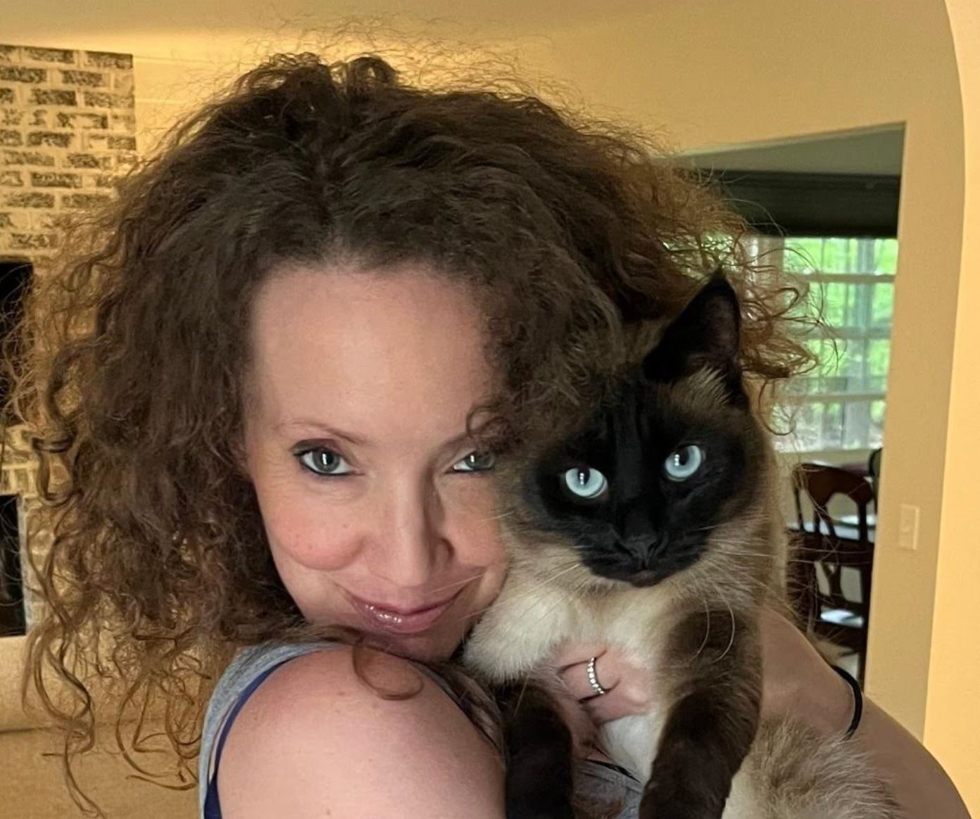 En person med krøllete hår som smiler mens han holder en siameserkatt tett inntil ansiktet innendørs