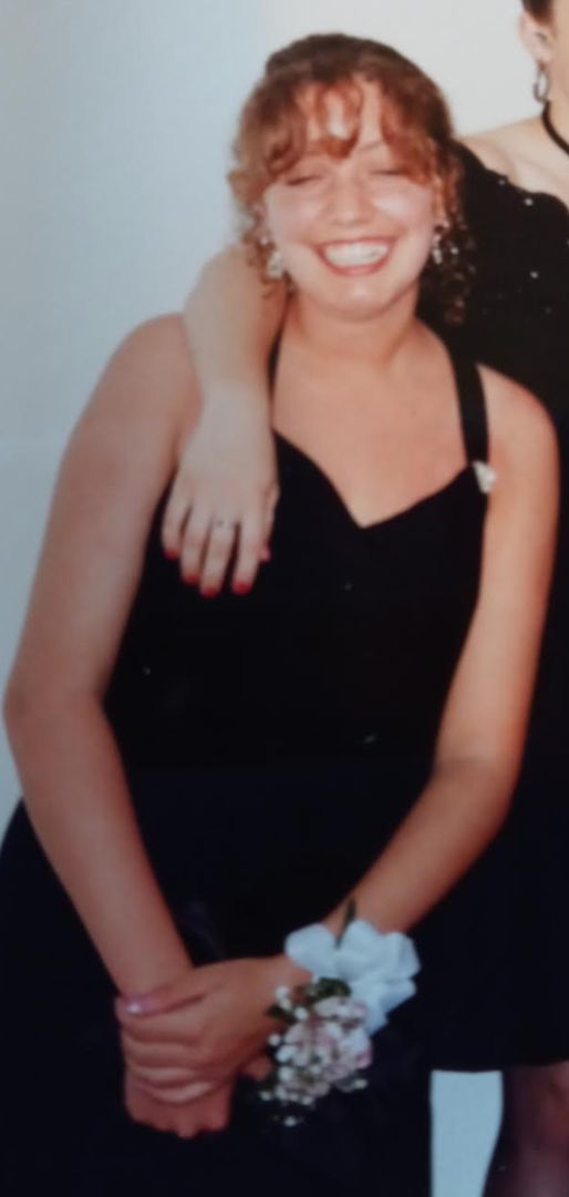 En ung kvinne i en svart kjole står og smiler og holder en liten bukett med en hvit blomst. Det er en arm over skulderen hennes