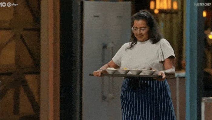 En kokk bærer et brett med boller i en kjøkkensetting, iført en hvit topp og stripete forkle