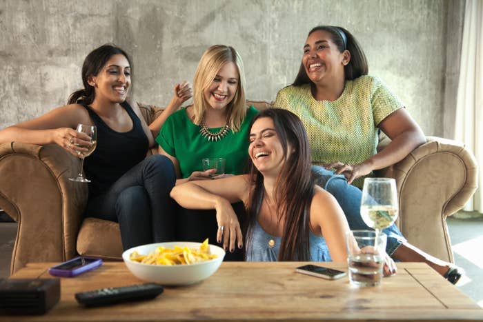 Fire kvinner ler og nyter drinker i en koselig stue, sittende på en sofa med snacks på bordet