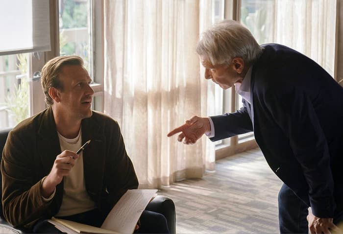 To personer i en stue; den ene sitter og holder et manus, mens den andre lener seg inn og peker. De virker engasjert i en diskusjon