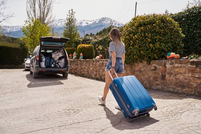 Person som trekker en koffert mot en bil lastet med bagger, plassert i et fjellrikt boligområde. Medfører forberedelse til tur eller ferie