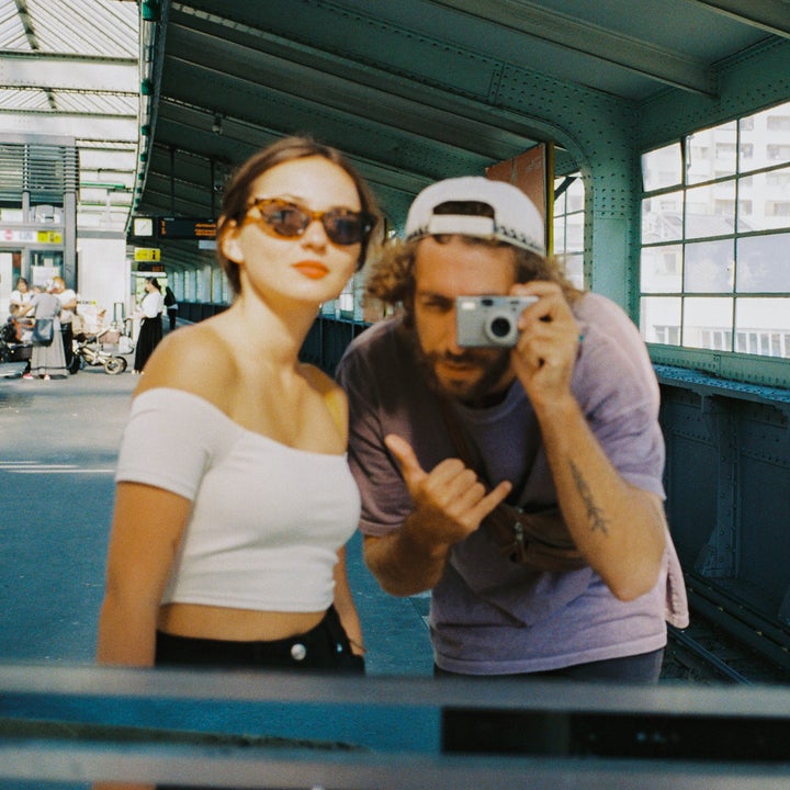 To personer på en togplattform tar en selfie i et speil. Den ene har på seg solbriller og en off-shoulder topp, den andre en caps og holder et kamera