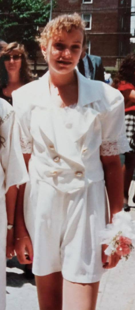 En person i et formelt hvitt antrekk med blomstertilbehør står utendørs; folk er i bakgrunnen